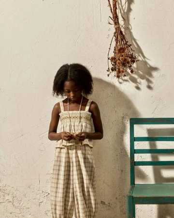 Young child in a plaid outfit standing beside a rustic teal chair against a textured beige wall with dried botanicals