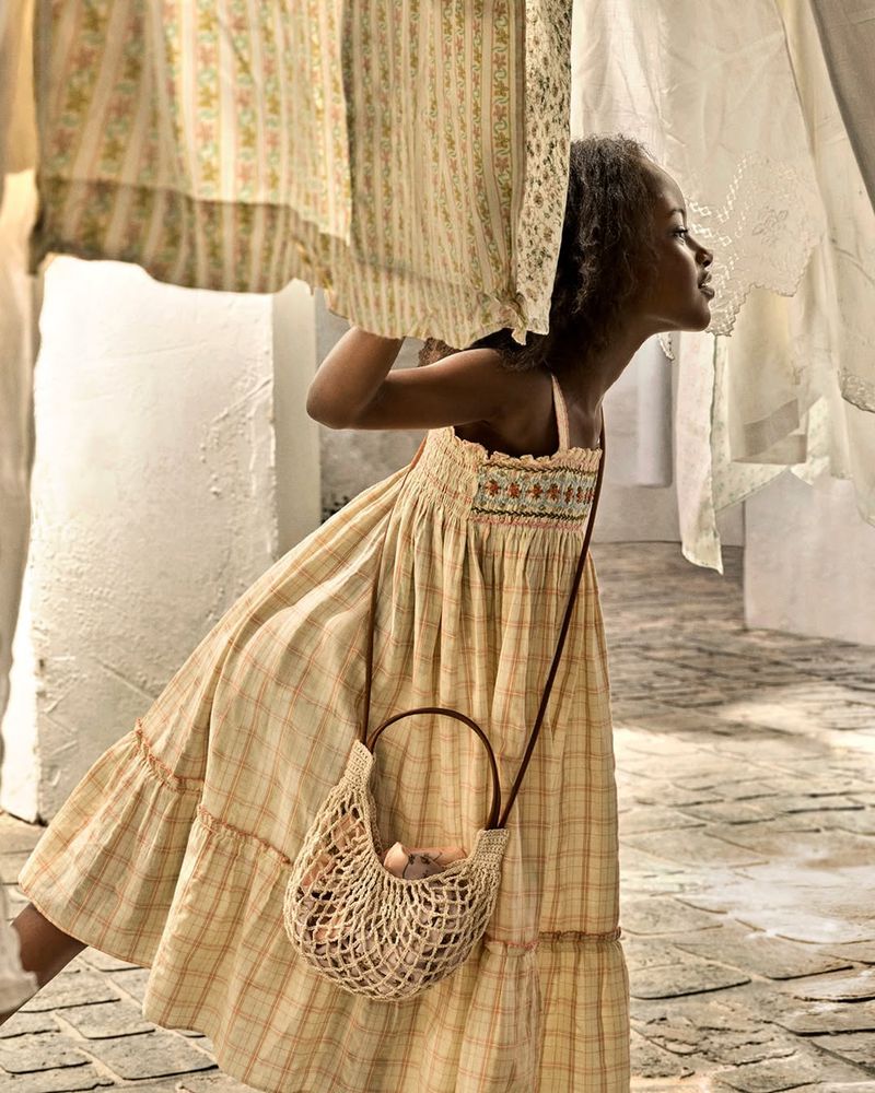 A young girl in a plaid sundress holding a woven mesh bag while walking outdoors past hanging fabrics.