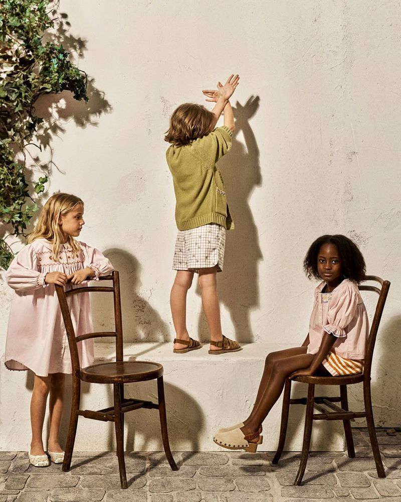 Three children posing outdoors against a textured white wall, two seated on wooden chairs and one standing