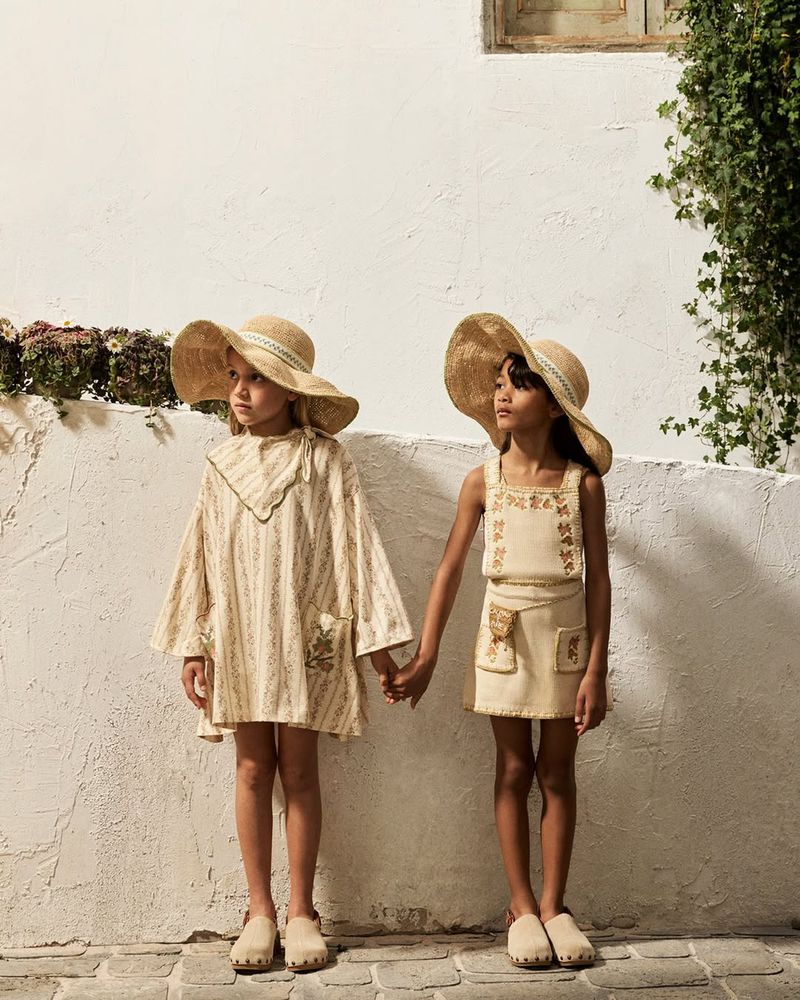 Two young girls holding hands in matching wide-brim straw hats and neutral outfits standing against a textured white wall.