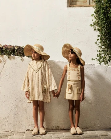 Two young girls holding hands in matching wide-brim straw hats and neutral outfits standing against a textured white wall.