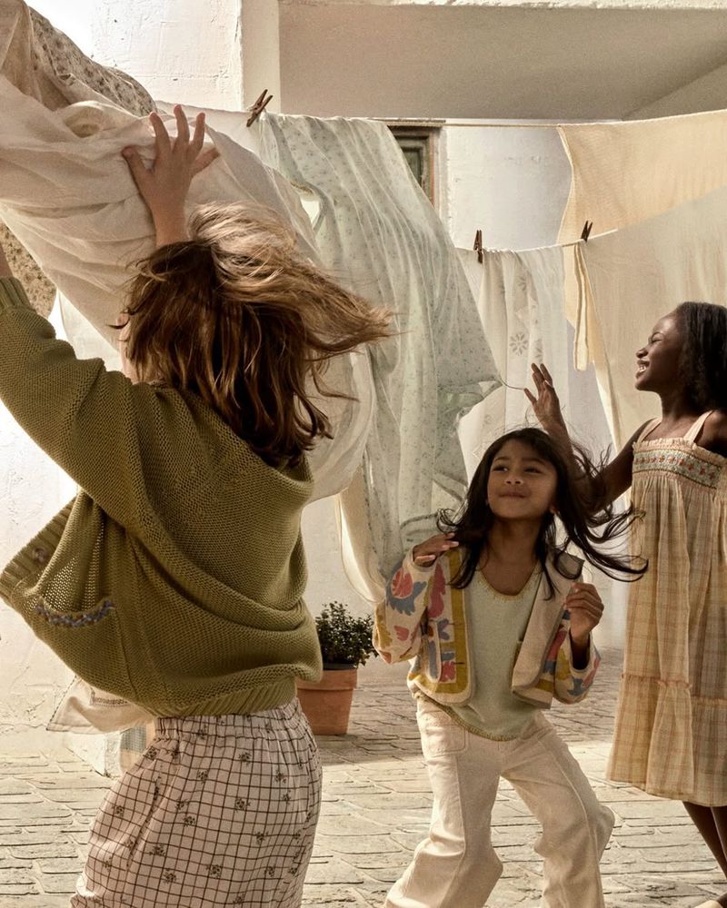 Three young girls in casual clothing play near hanging laundry outdoors on a sunny day, showcasing childhood, fashion, and lifestyle.
