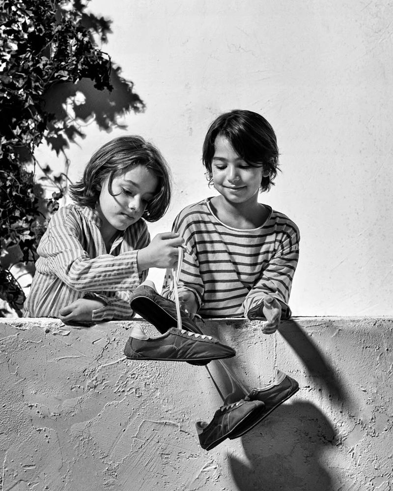 Two children leaning over a wall holding up a pair of sneakers by their laces in a black and white photograph.
