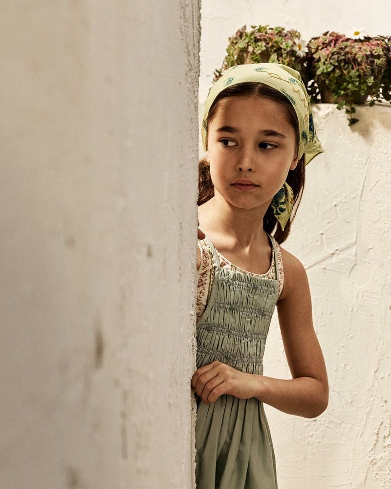 Young girl wearing a green headscarf and smocked dress peering around a white wall outdoors for Zara Kids