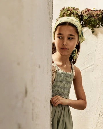 Young girl wearing a green headscarf and smocked dress peering around a white wall outdoors for Zara Kids