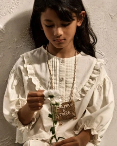 Young child wearing a cream ruffled blouse holding a white flower against a textured neutral wall for Zara Kids