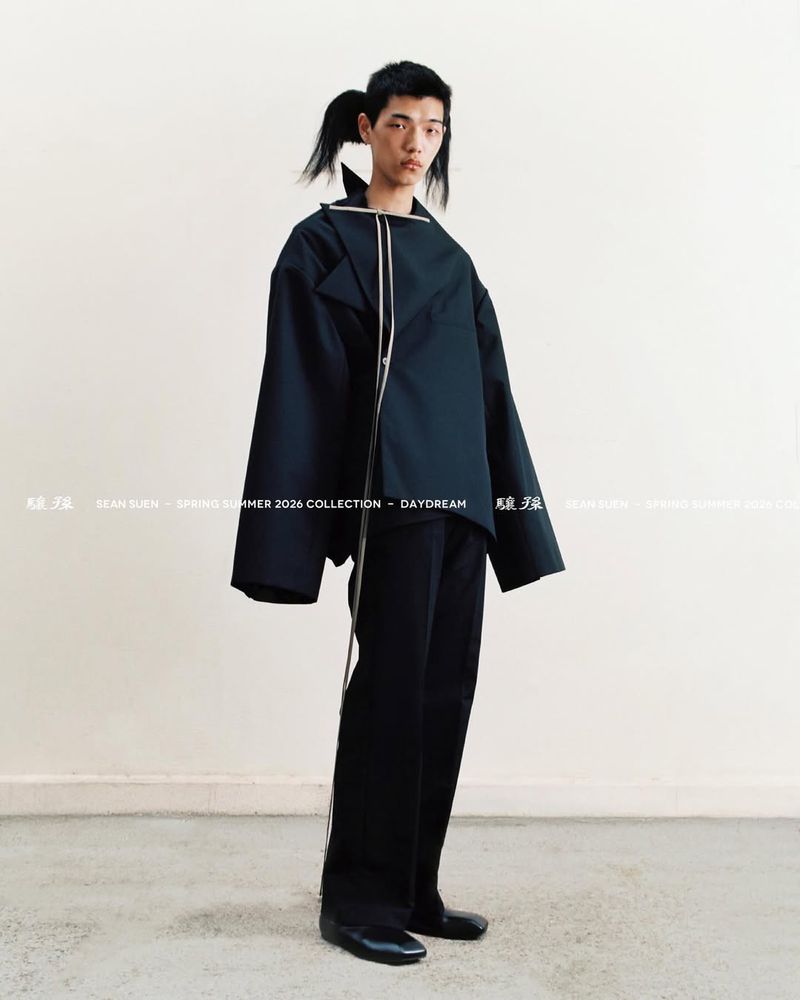 Fashion-forward adult male model showcases an oversized black jacket with white trim, paired with wide-leg pants in a minimalist studio.
