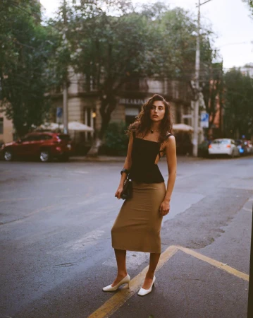 Model Paola Manes standing on an urban street wearing a black top and tan skirt for Reformation Spring 2026