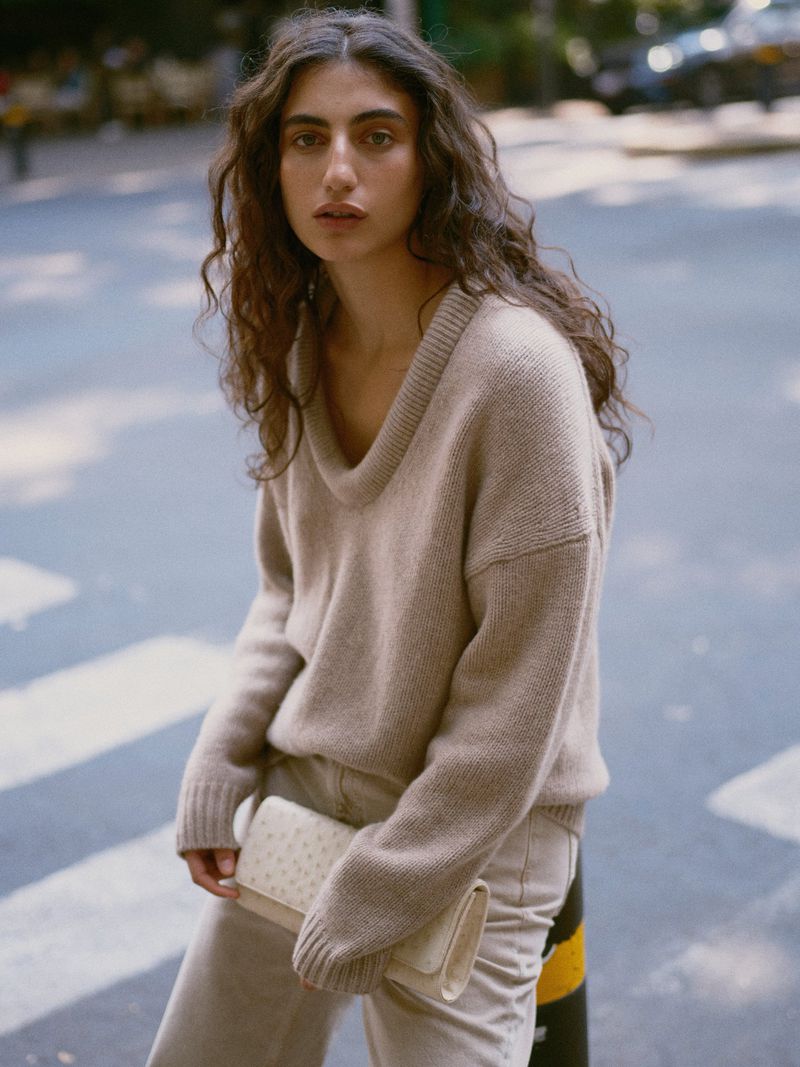 Model with long curly dark hair wearing a beige V-neck sweater holding a cream clutch on a city street