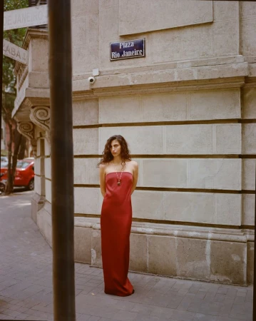 Model wearing a long red strapless dress standing on a sidewalk in front of a stone building exterior with a street sign