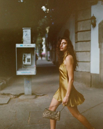 Model in gold satin mini dress walking down a city street past a public telephone booth holding an animal print handbag
