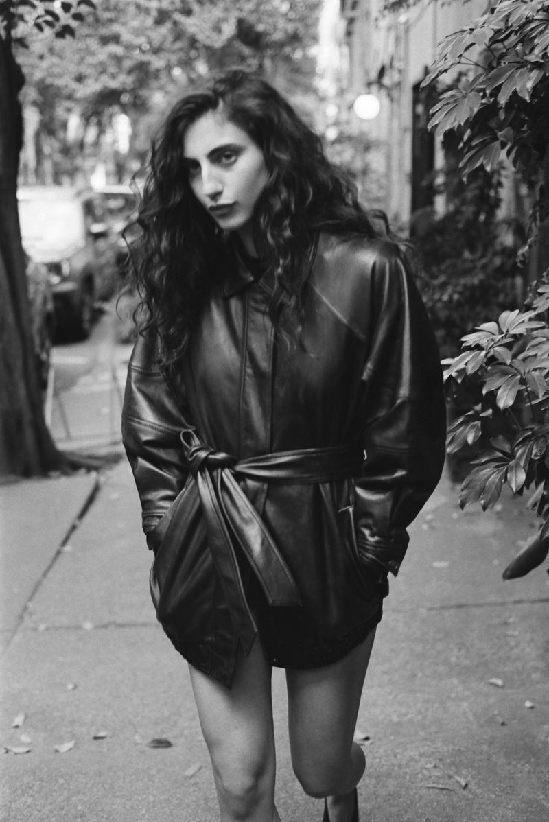 Model with long dark wavy hair wearing a belted leather jacket walking on a city sidewalk