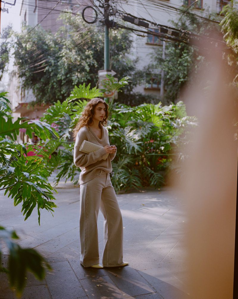 Model in neutral sweater and wide trousers holding a clutch bag amidst lush green foliage