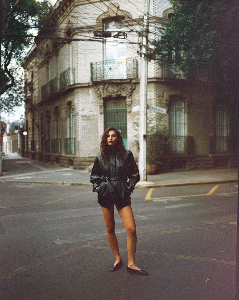 Model standing on a city street corner wearing a dark belted leather jacket and flat shoes.