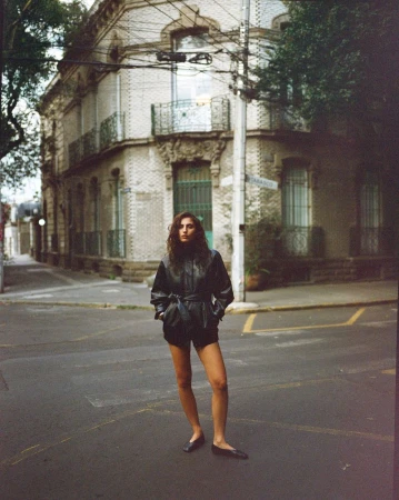 Model standing on a city street corner wearing a dark belted leather jacket and flat shoes.