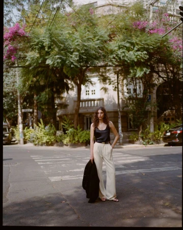 Woman wearing a black silk top and white trousers standing on a sunny street holding a dark jacket