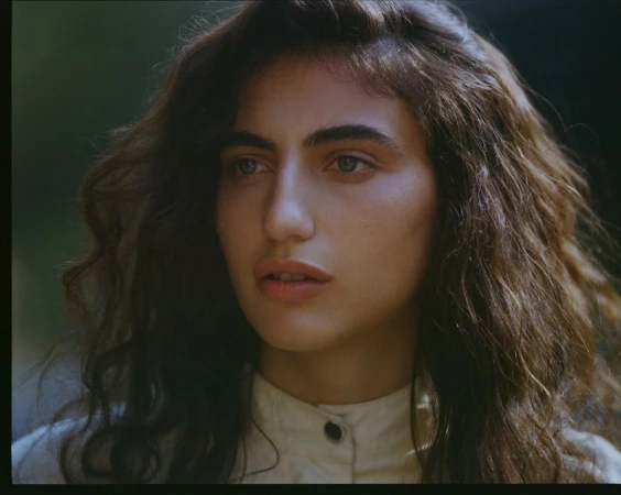 Close-up portrait of a woman with long dark wavy hair wearing an off-white buttoned top outdoors.