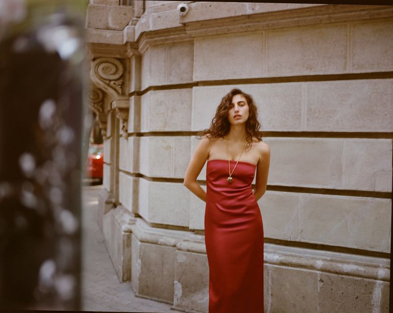 Model wearing a strapless red midi dress and long gold pendant necklace posing against a classical stone building exterior.