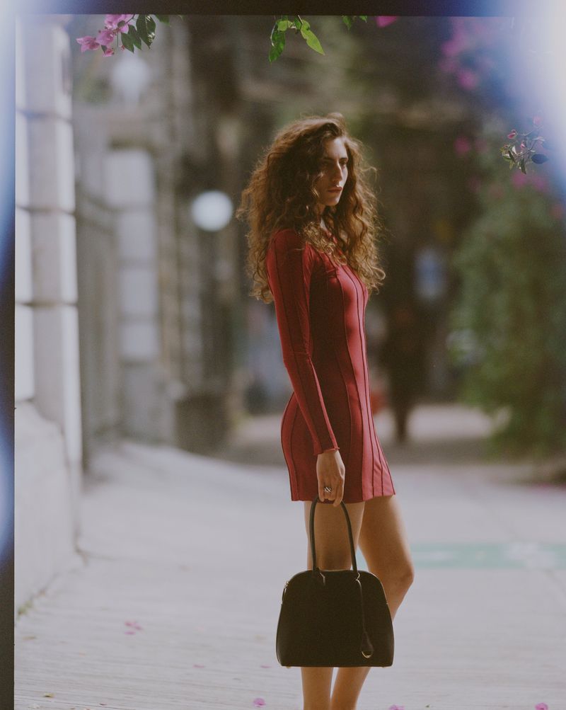 Woman with long curly hair wearing a fitted red dress holding a black handbag outdoors on a city sidewalk