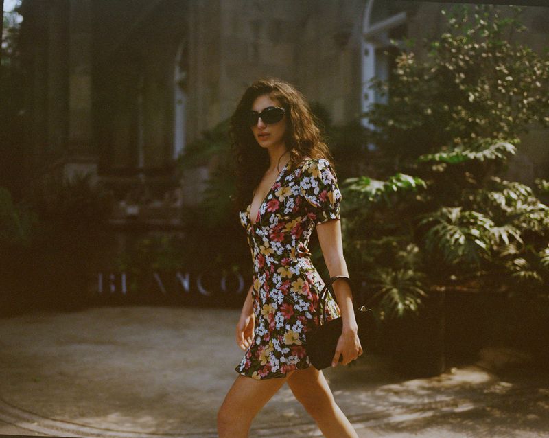 Model wearing a floral print mini dress and sunglasses holding a black handbag outdoors