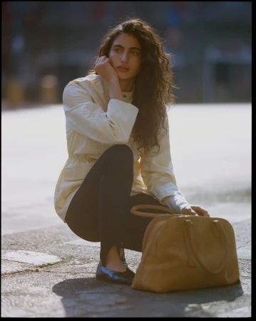 Model with long curly hair crouching outdoors wearing a cream jacket, dark stirrup leggings, and holding a tan leather handbag