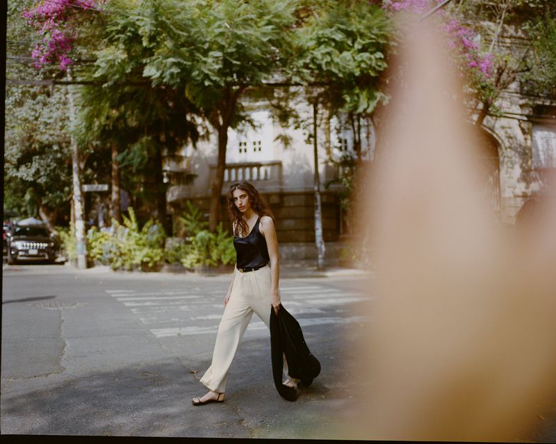 Model wearing a black top and light trousers walking on a street, partially obscured in the foreground