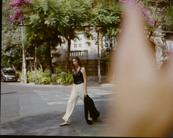 Model wearing a black top and light trousers walking on a street, partially obscured in the foreground