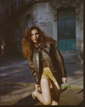 Model with long curly brown hair wearing a dark leather jacket and shorts outdoors on a street.