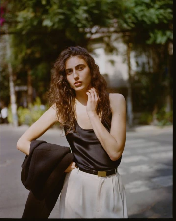 Model in a black silk top and light trousers holding a dark jacket outdoors in a street setting for Reformation Spring 2026.