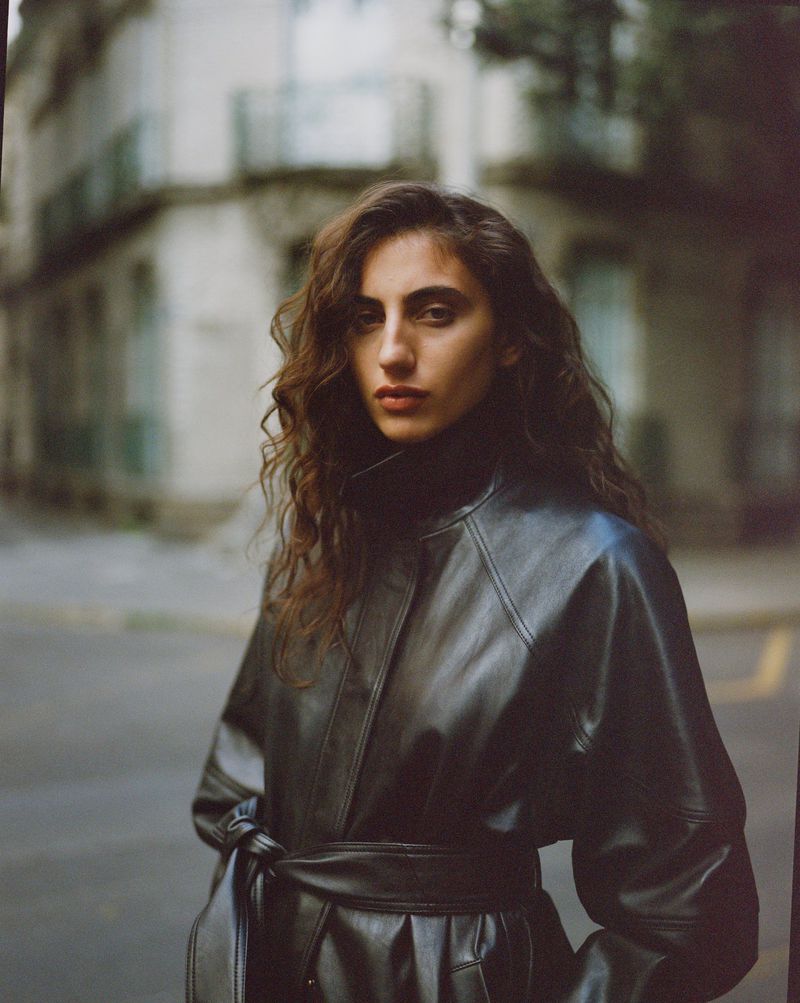 Model wearing a black leather belted coat standing on a city street for Reformation Spring 2026.