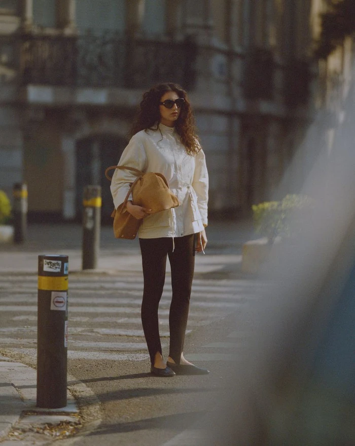 Model standing on a city street wearing a white jacket, dark leggings, sunglasses, and holding a tan handbag.