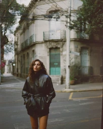 Model wearing a dark belted leather jacket standing on an urban street corner in front of a classic building.