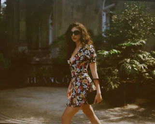 Model wearing a floral print mini dress and sunglasses holding a black handbag outdoors