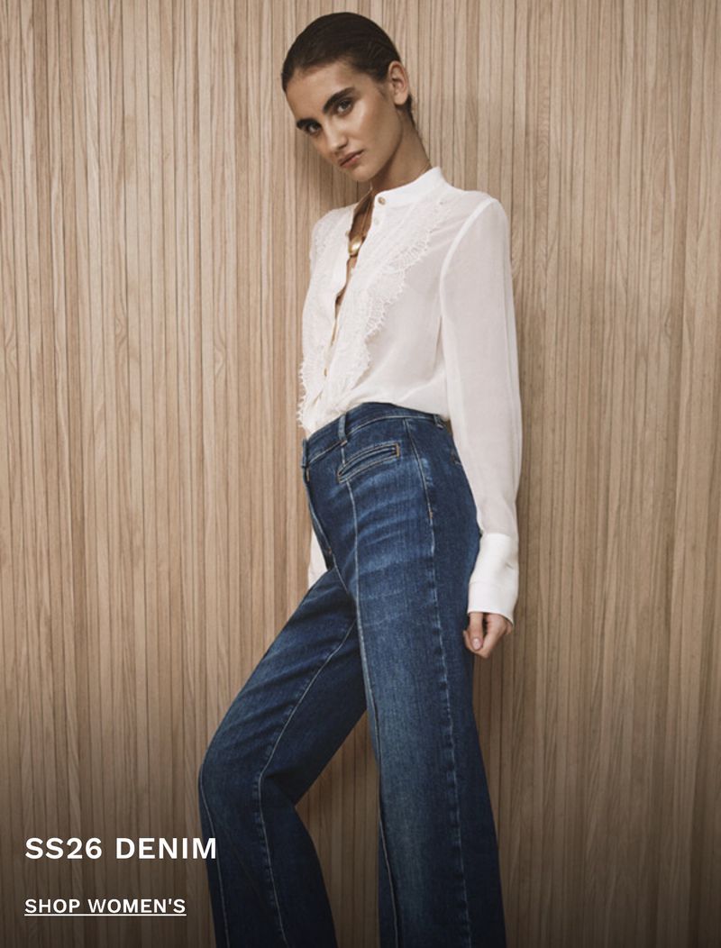 Model wearing a white blouse and dark blue denim jeans standing against a vertical wood panel wall for Reiss Denim S/S 26.