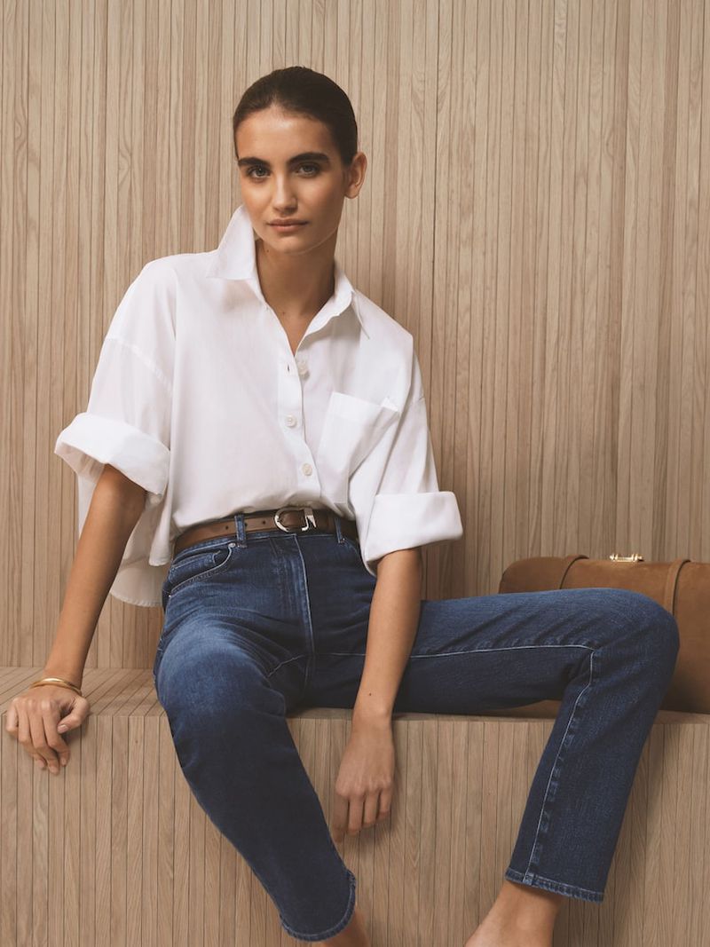 A fashionable adult female sits indoors wearing a white button-down shirt, blue jeans, and a brown belt in a medium shot.