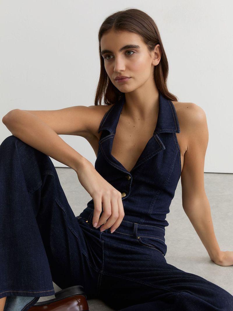 Model seated wearing a dark denim halter top and matching trousers against a plain white studio wall.