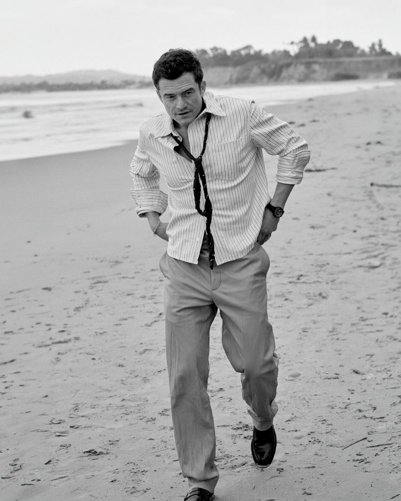 Actor Orlando Bloom walking along a sandy beach wearing a striped button-down shirt, loose tie, and trousers in black and white.