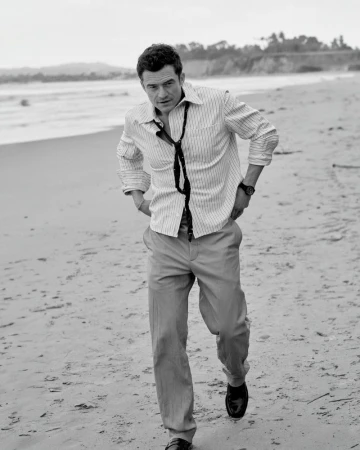 Actor Orlando Bloom walking along a sandy beach wearing a striped button-down shirt, loose tie, and trousers in black and white.