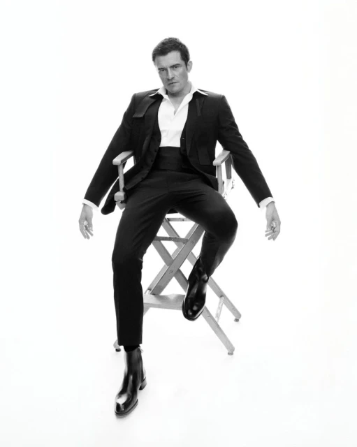 Black and white portrait of a man in a suit sitting on a director's chair in a photo studio against a bright white backdrop.
