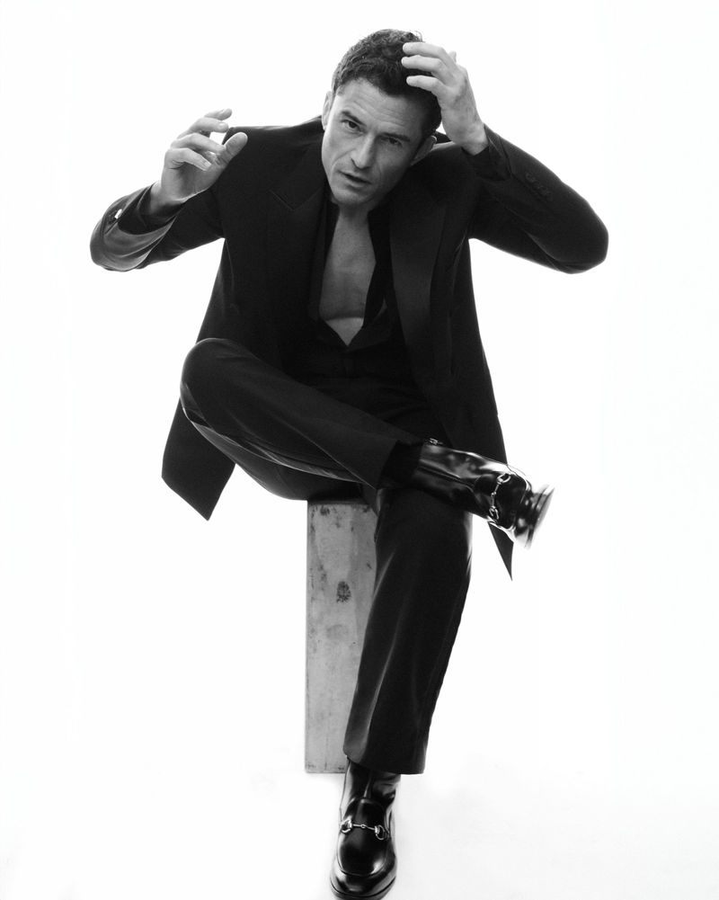 Black and white portrait of a man sitting on a block in a relaxed pose, wearing a suit with an open shirt, looking at the camera.