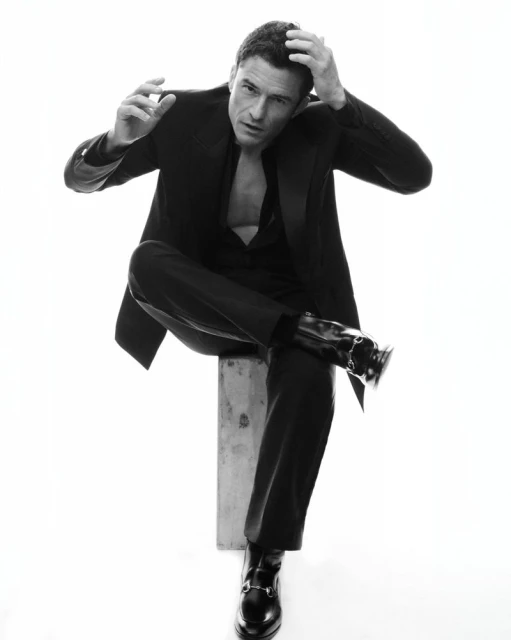 Black and white portrait of a man sitting on a block in a relaxed pose, wearing a suit with an open shirt, looking at the camera.