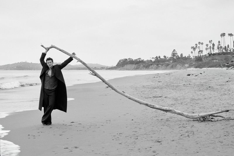 Man stands on beach holding a large branch with ocean and sky in background, styled in black and white.
