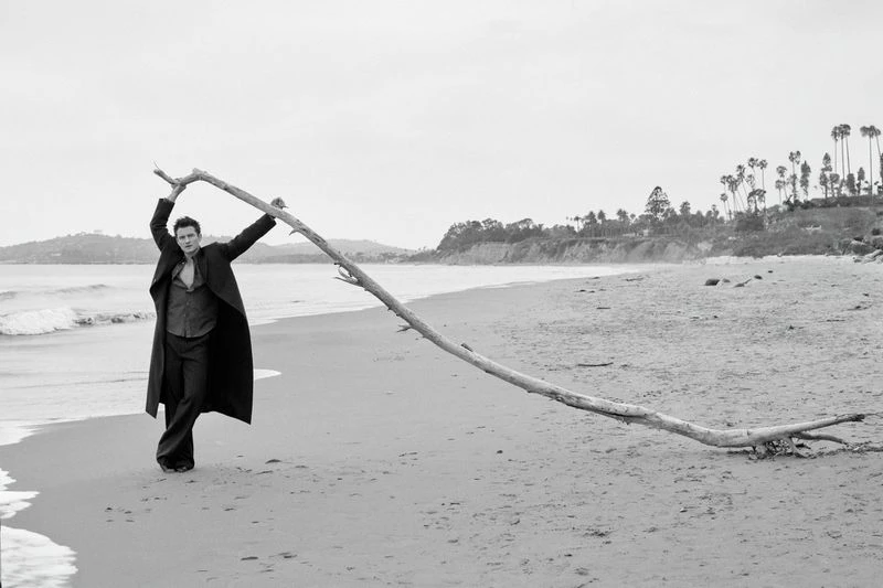 Man stands on beach holding a large branch with ocean and sky in background, styled in black and white.