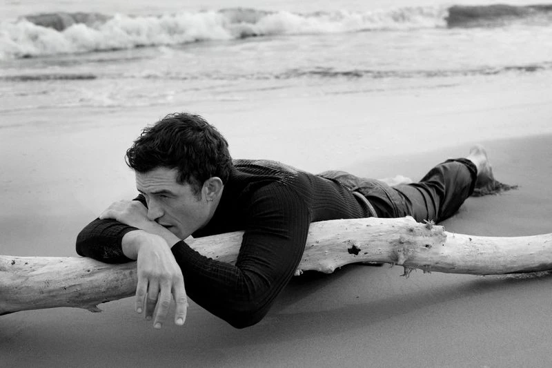 Orlando Bloom lounges on driftwood at the beach, eyes averted, his long-sleeved black shirt contrasting with the monochrome seascape.