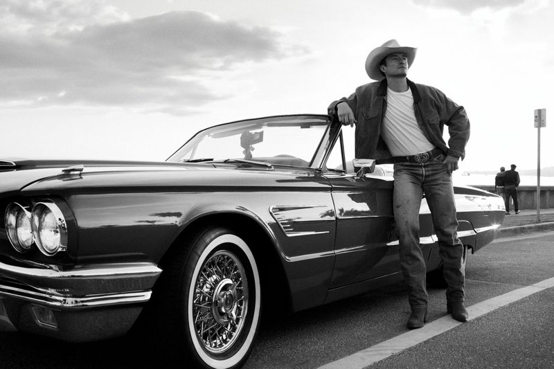 A man wearing a cowboy hat leans on a vintage convertible car in a black and white outdoor shot, showcasing classic automotive style.