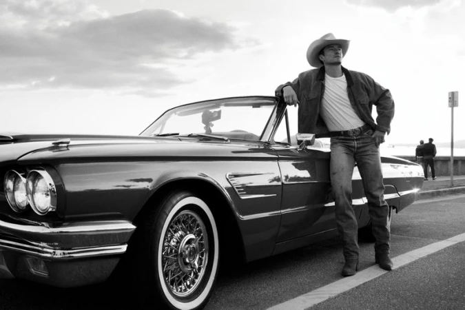 A man wearing a cowboy hat leans on a vintage convertible car in a black and white outdoor shot, showcasing classic automotive style.