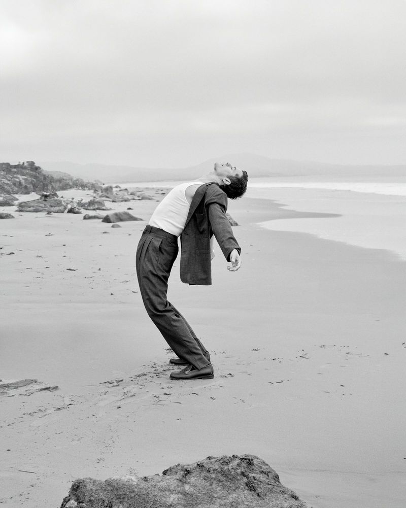 A man in suit pants and a white tank top arches backward on a sandy beach, with rocks, sand, and water in the black-and-white photograph.