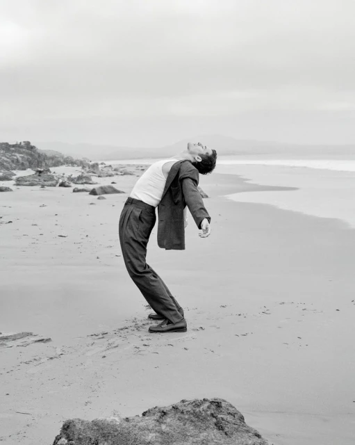 A man in suit pants and a white tank top arches backward on a sandy beach, with rocks, sand, and water in the black-and-white photograph.