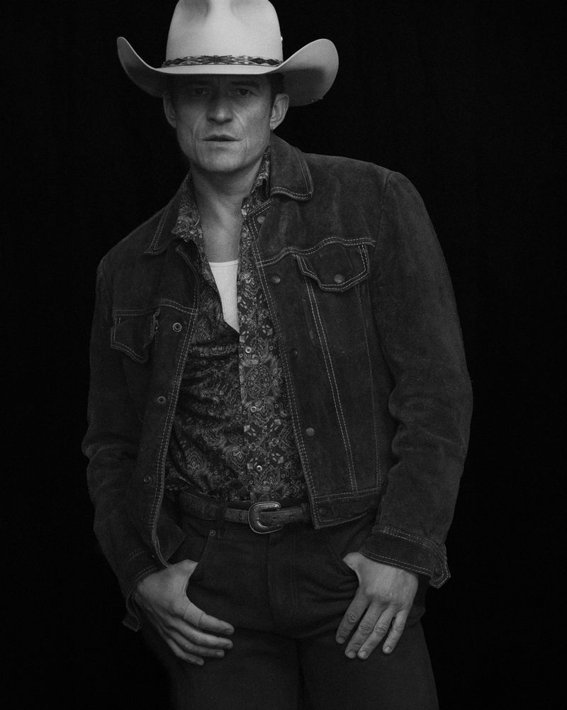 Orlando Bloom wearing a cowboy hat, unbuttoned patterned shirt, and dark jacket in a black-and-white studio portrait.