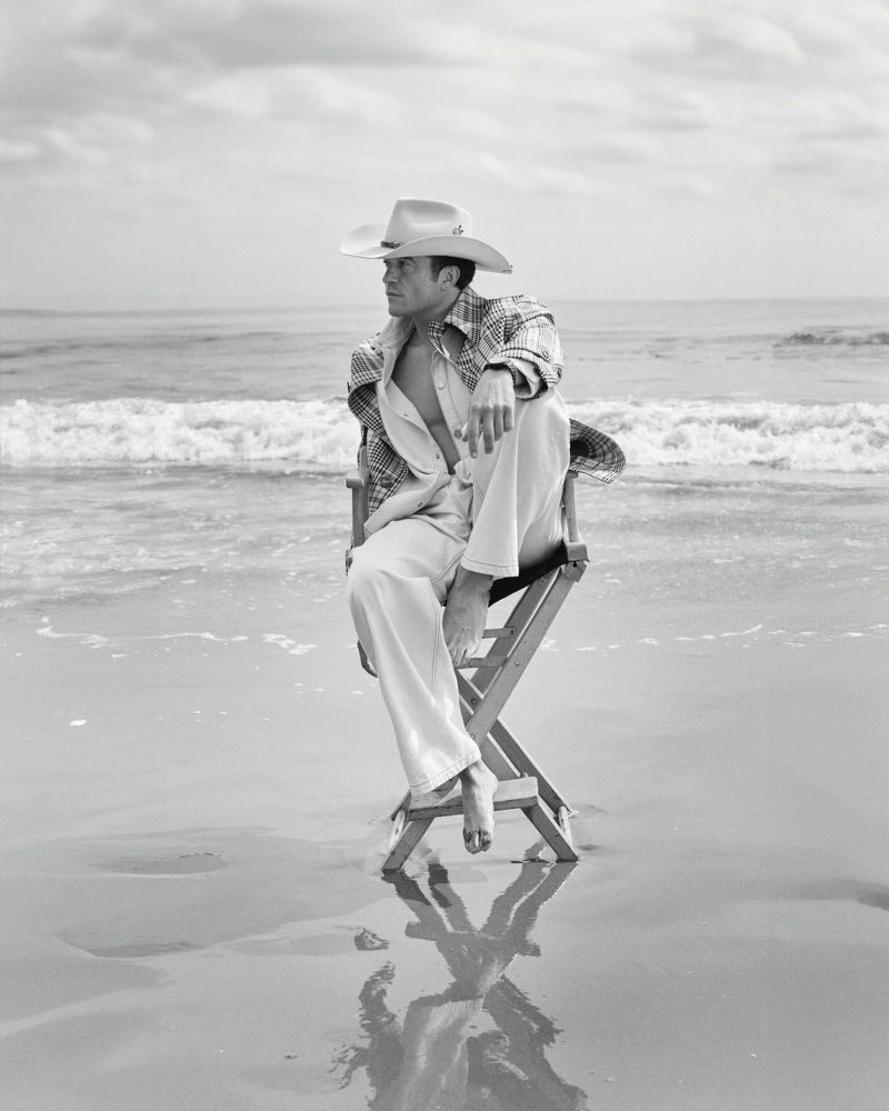 Orlando Bloom barefoot in a cowboy hat sitting on a director chair in shallow ocean surf on a sandy beach in black and white.